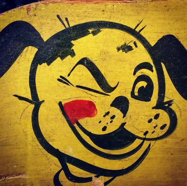 The original hand-painted Pet Pantry winking dog, black line and one red tongue on a scuffed yellow board
